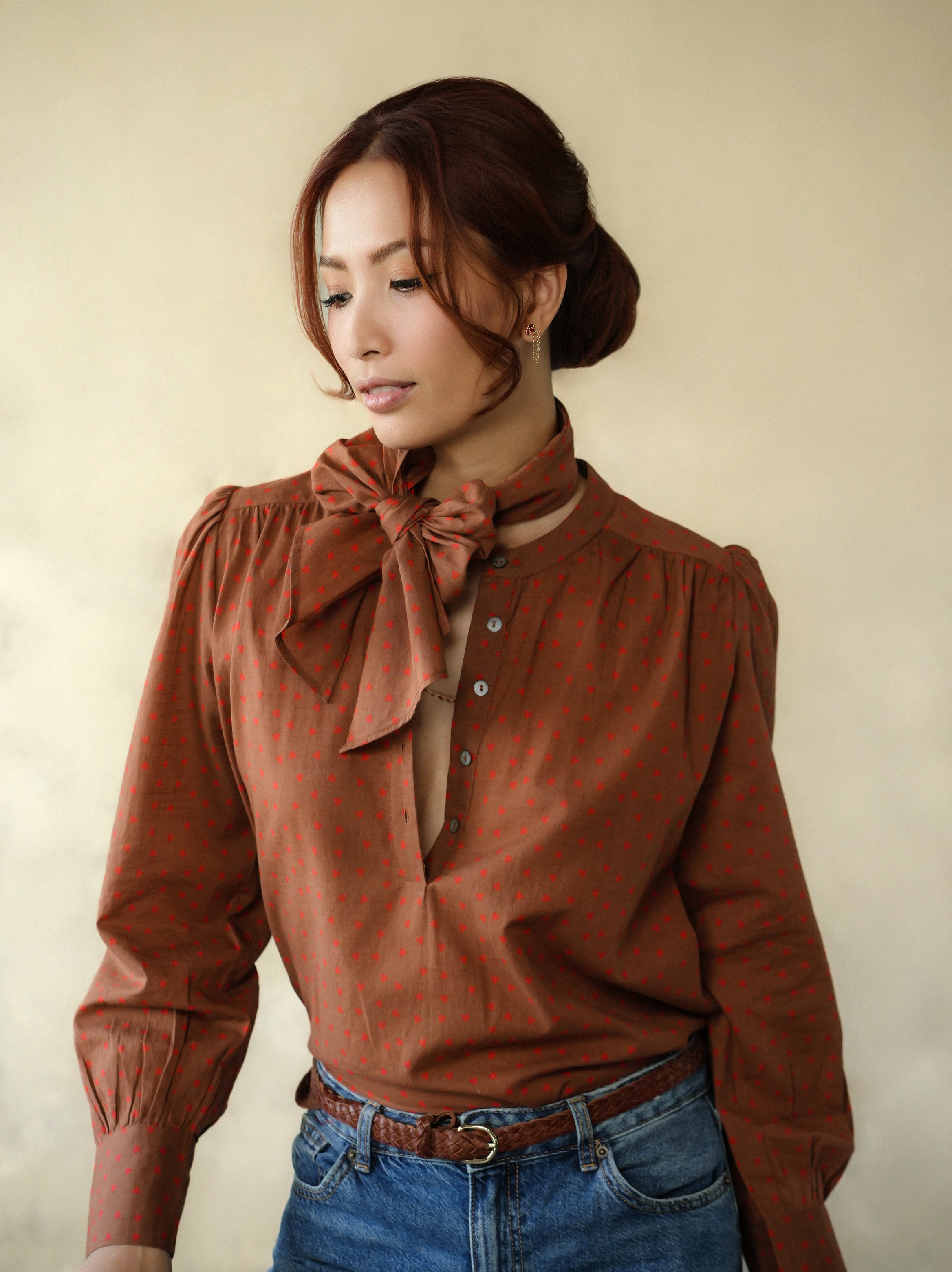 Woman wearing a brown blouse with a bow tie and blue jeans against a beige background
